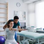 Young male physiotherapist exercising with young woman patient on ball in a physic room